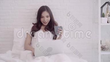 亚洲妇女使用电脑或笔记本电脑，拿着一杯温暖的咖啡或茶，躺在床上，当她在家里的卧室放松。